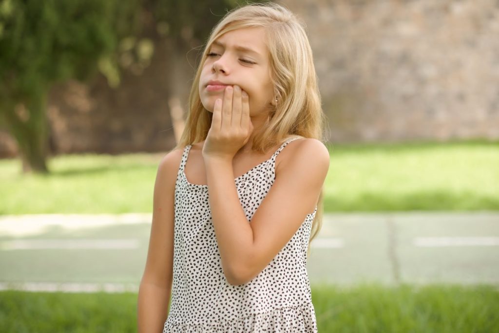 Child with toothache outdoors, holding cheek.